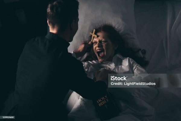 male exorcist with bible and cross standing over demoniacal screaming girl in bed Exorcism Stock Photo